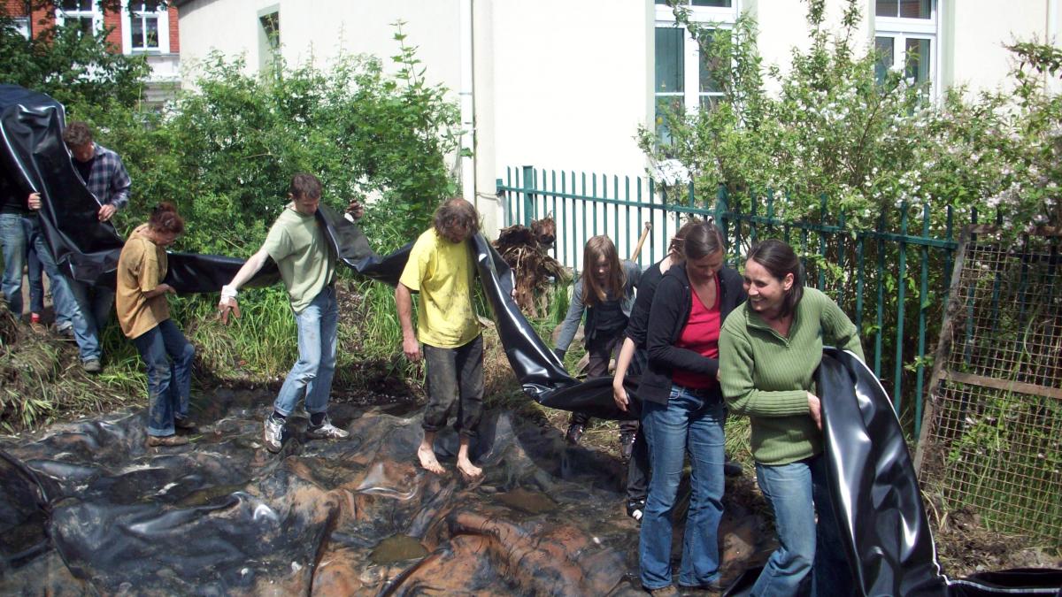 Mehrere Menschen tragen eine große Folie um einen Teich damit auszulegen